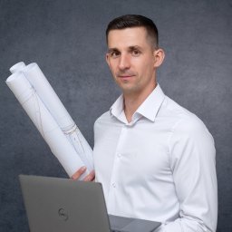 GEOGROUP BŁAŻEJ OSTROWSKI - Młody mężczyzna w białej koszuli trzyma laptopa i zwinięte plany budowlane na tle szarej ściany. Uśmiechnięty, patrzy wprost.