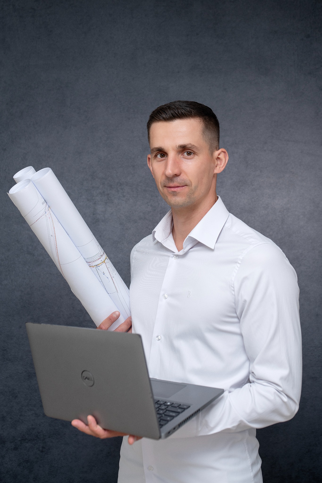 Młody mężczyzna w białej koszuli trzyma laptopa i zwinięte plany budowlane na tle szarej ściany. Uśmiechnięty, patrzy wprost.