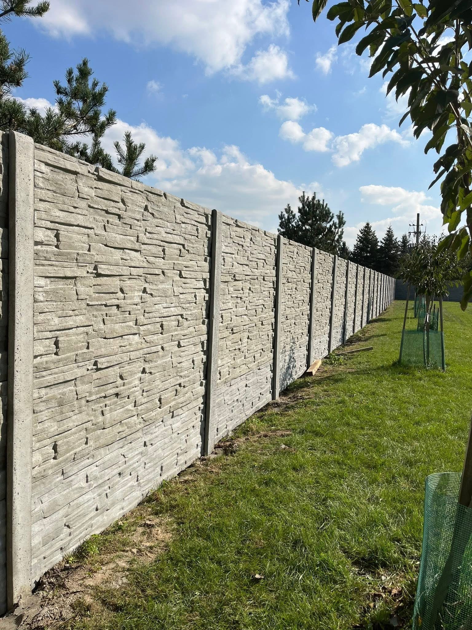 Długi, szary mur z betonu imitującego kamień, oddzielający zielony trawnik od dalszego terenu. Widok z perspektywy, błękitne niebo z chmurami w tle.