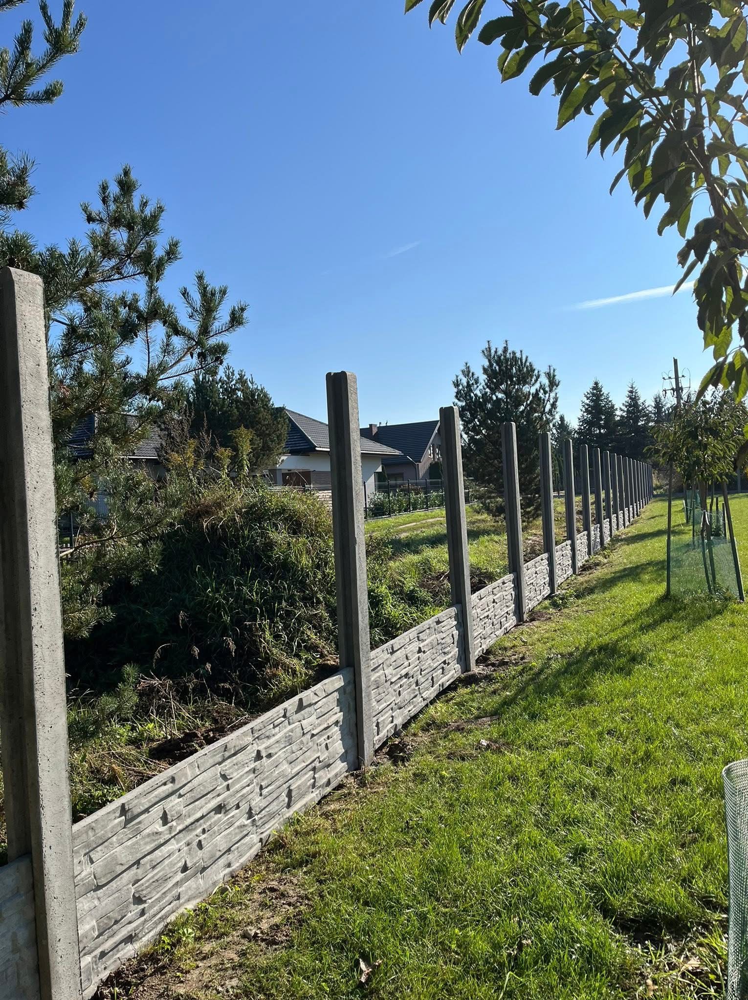 Nowoczesne ogrodzenie betonowe imitujące kamień, z widocznymi słupkami, na tle zielonego trawnika i błękitnego nieba. Ujęcie z perspektywy ziemi.