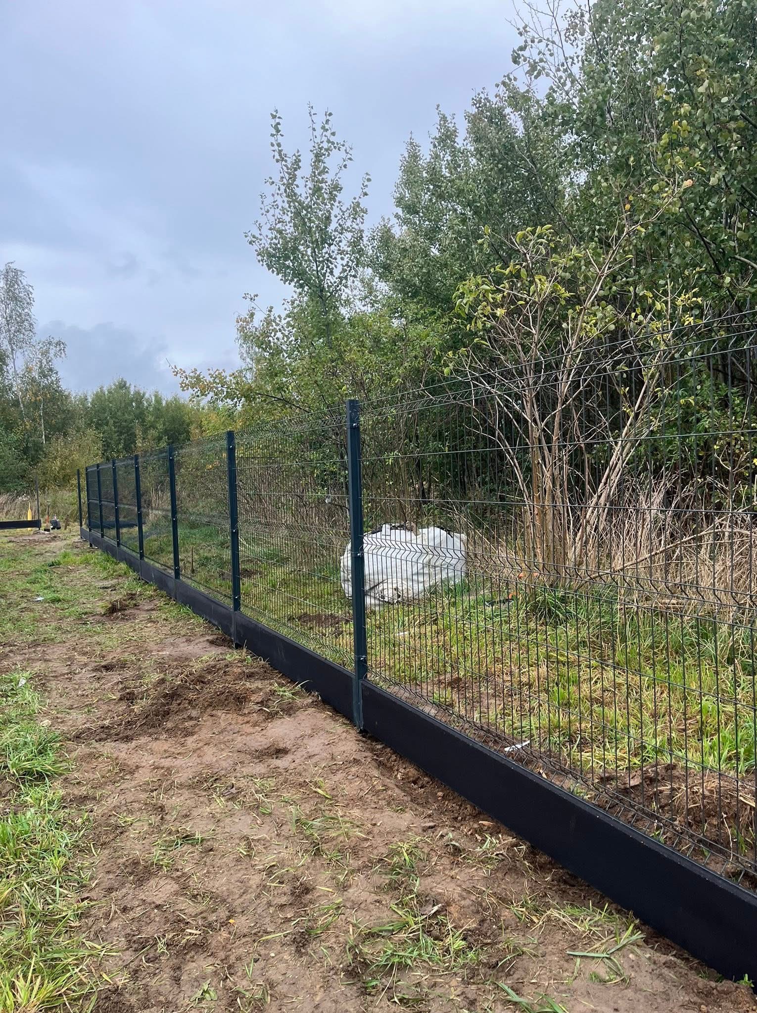 Nowoczesne ogrodzenie panelowe z podmurówką w kolorze antracytu, na tle zieleni drzew. Widoczne worki z materiałem budowlanym. Ujęcie z poziomu gruntu.
