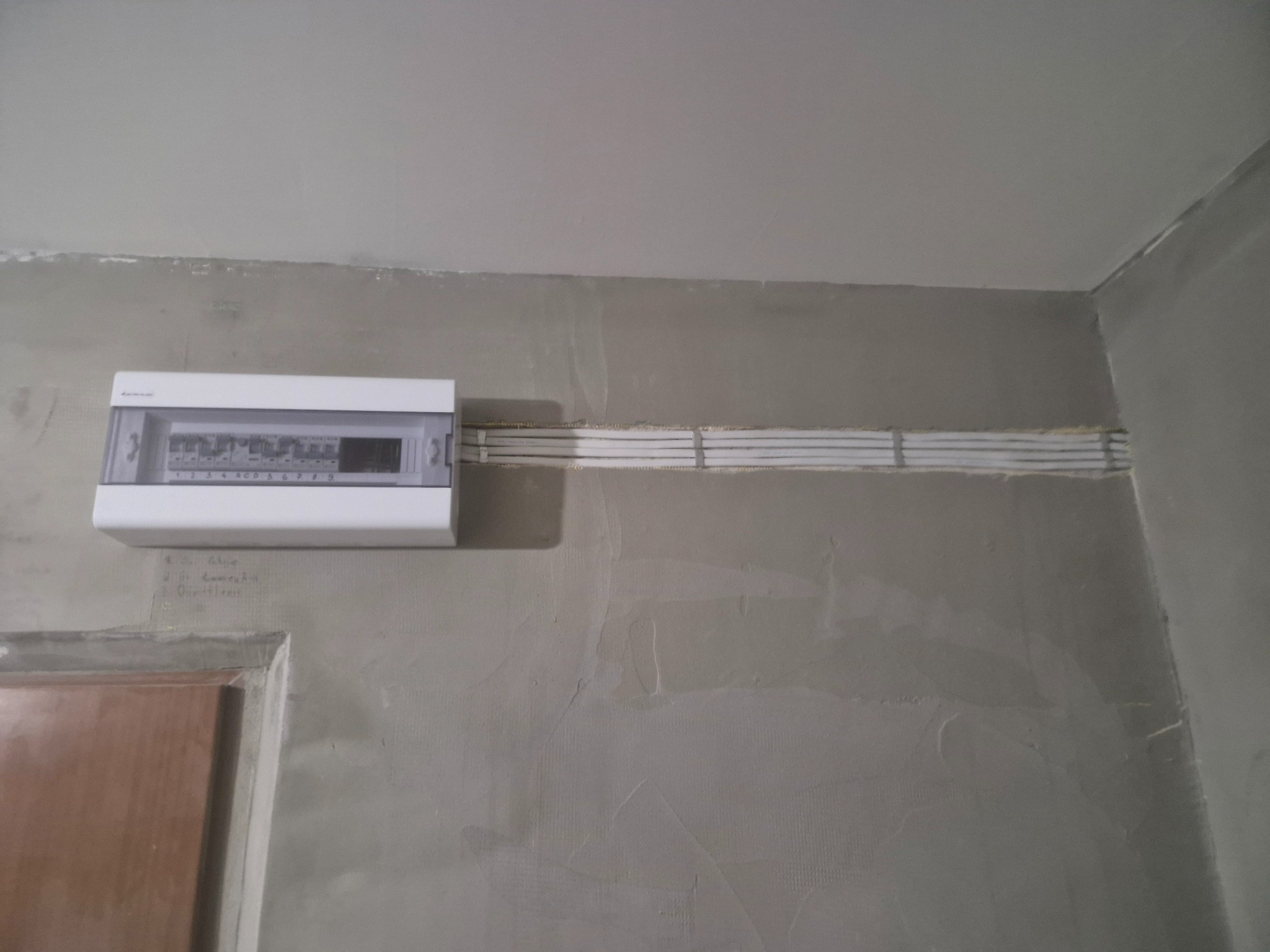 Biała skrzynka rozdzielcza z przewodami na szarej, surowej ścianie w trakcie wykończenia. Widoczne oznaczenia na ścianie i fragment ościeżnicy drzwi.