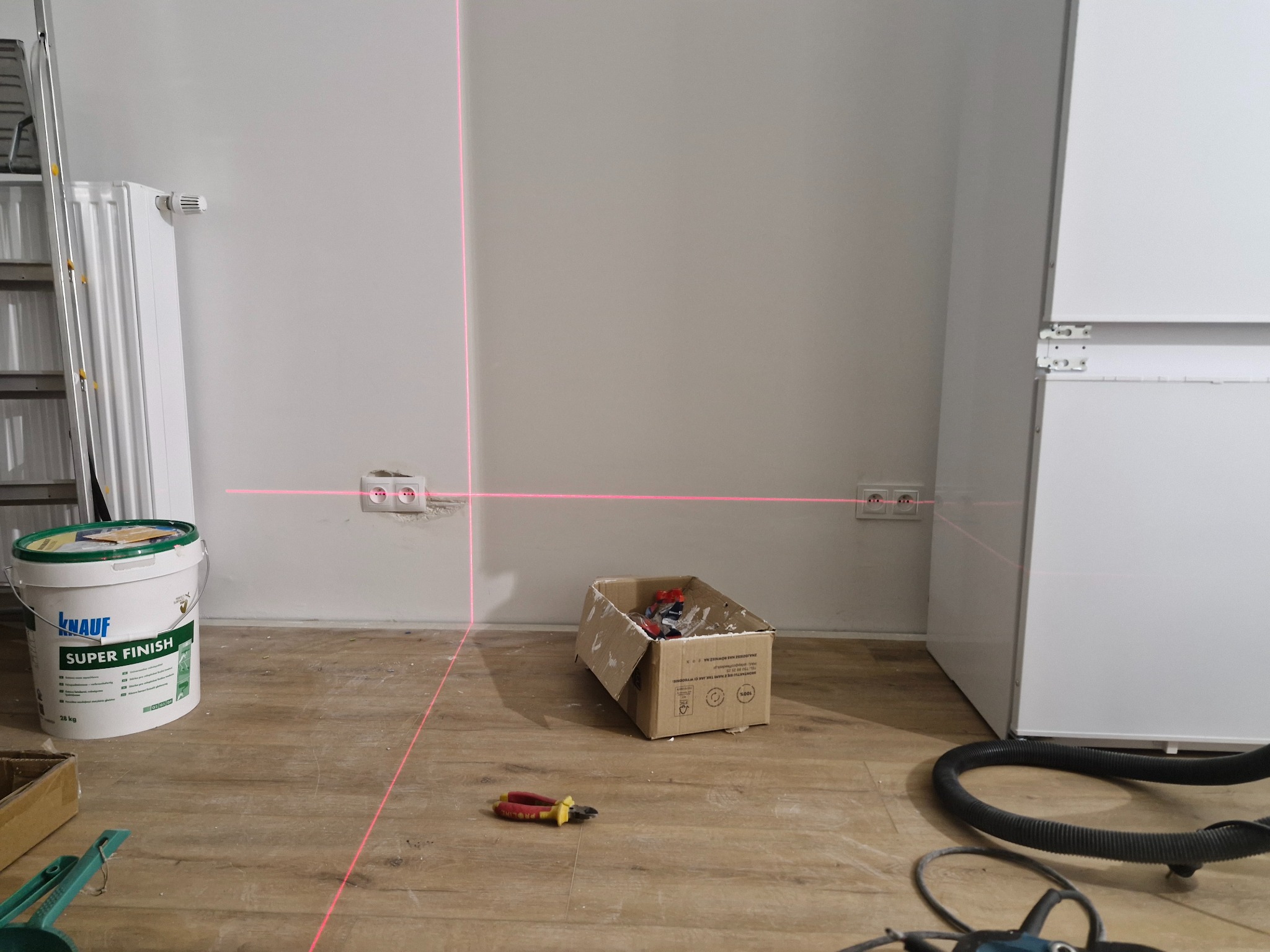 Poziomica laserowa wyznacza linie na ścianie z gniazdkami elektrycznymi. Na podłodze wiadro z gładzią, karton z narzędziami i przewód odkurzacza.