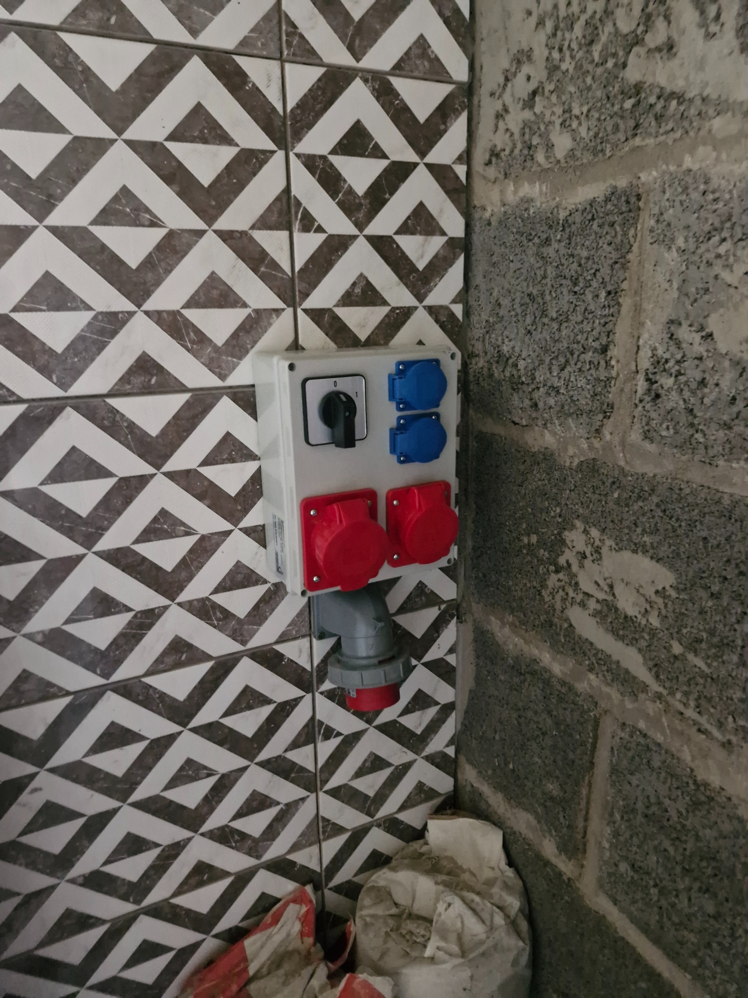 Zamontowana, natynkowa skrzynka elektryczna z gniazdami siłowymi (czerwone) i standardowymi (niebieskie) na ścianie z wzorzystych płytek i surowego betonu.