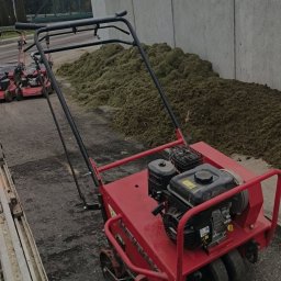 GREEN CRAFT - Czerwony aerator spalinowy na tle betonowego muru i sterty skoszonej trawy. W tle widoczny inny sprzęt ogrodniczy i drzewa.