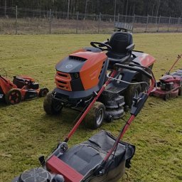 GREEN CRAFT - Różne kosiarki, w tym pomarańczowy traktor, stoją na trawniku przed lasem. Widoczne ślady użytkowania na maszynach. Sprzęt do koszenia trawy.