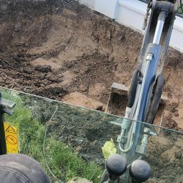 GREEN CRAFT - Wykop pod fundament ogrodzenia wykonany minikoparką, widok z kabiny operatora. Świeżo naruszona ziemia przy białym, ozdobnym ogrodzeniu.