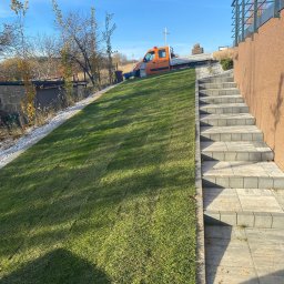 GREEN CRAFT - Świeżo założony trawnik na skarpie, z równo ułożonymi płytami schodowymi i pomarańczowym samochodem dostawczym w tle, w słoneczny dzień.