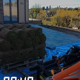 GREEN CRAFT - Paleta z rolkami trawy na tle nowo ułożonej kostki brukowej i budynku z numerem 12A, widziana z perspektywy maszyny budowlanej w słoneczny dzień.