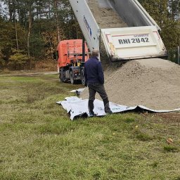 GREEN CRAFT - Ciężarówka wyładowuje piasek na trawniku, mężczyzna obserwuje proces. Widoczny las w tle. Wyładunek piasku na przygotowaną plandekę.