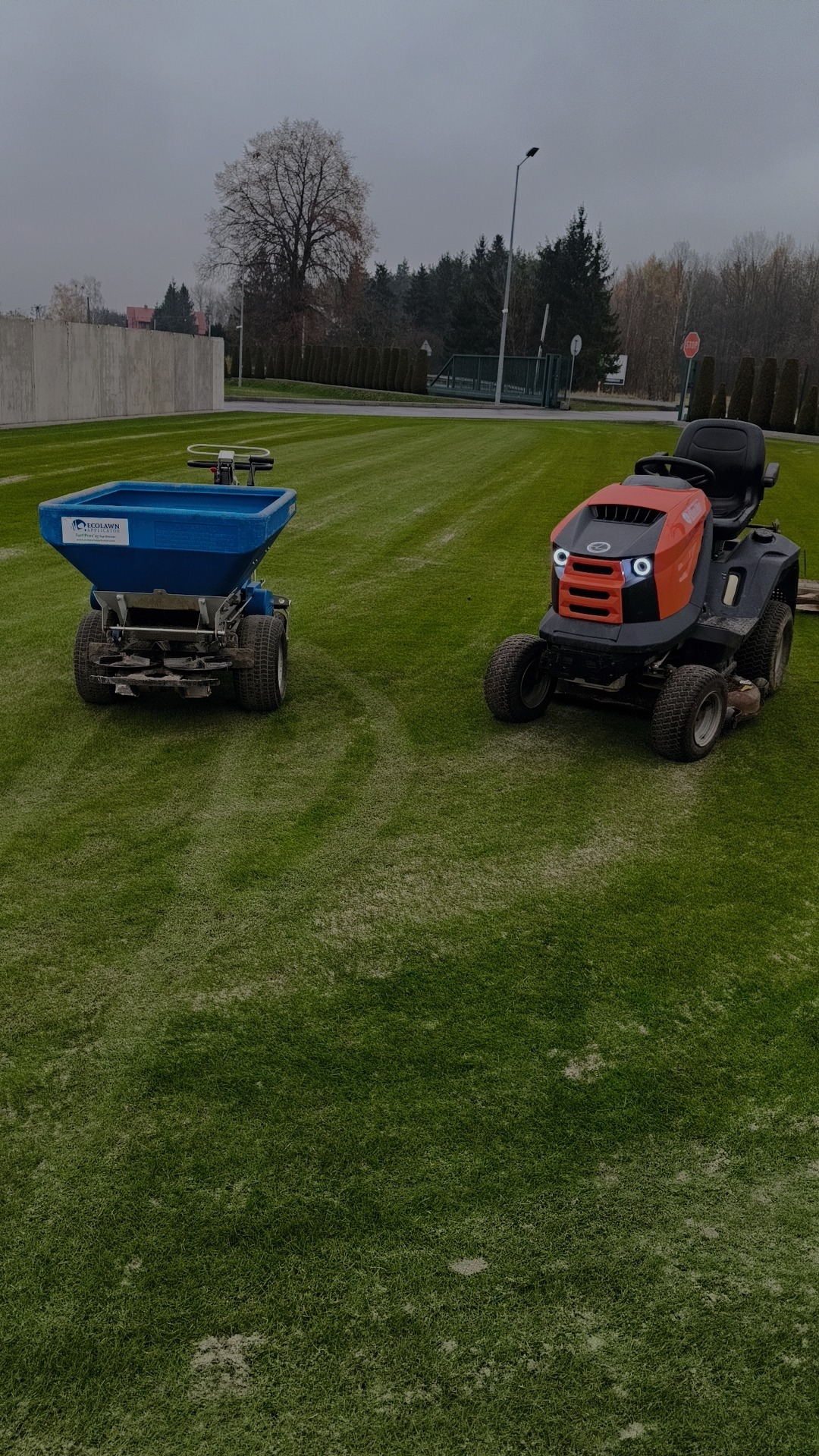 Traktor-kosiarka i rozsiewacz na trawniku po nawożeniu. Widoczne ślady nawozu. Sprzęt ogrodniczy na tle zieleni i zabudowań.