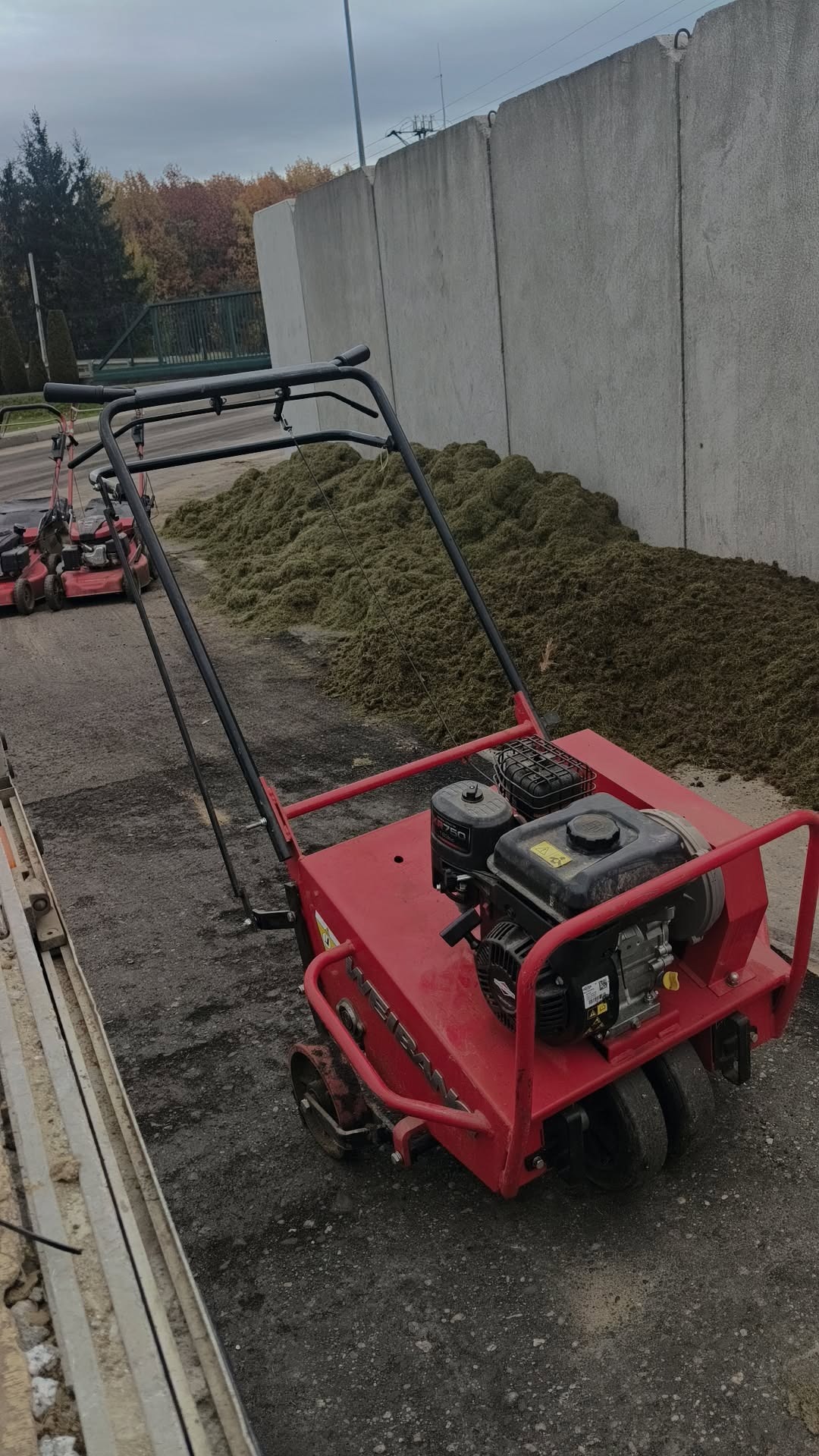 Czerwony aerator spalinowy na tle betonowego muru i sterty skoszonej trawy. W tle widoczny inny sprzęt ogrodniczy i drzewa.