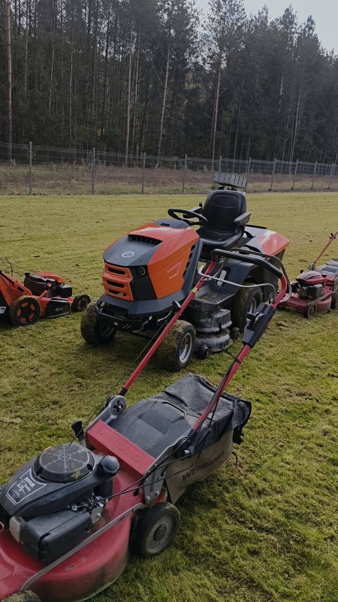 Różne kosiarki, w tym pomarańczowy traktor, stoją na trawniku przed lasem. Widoczne ślady użytkowania na maszynach. Sprzęt do koszenia trawy.