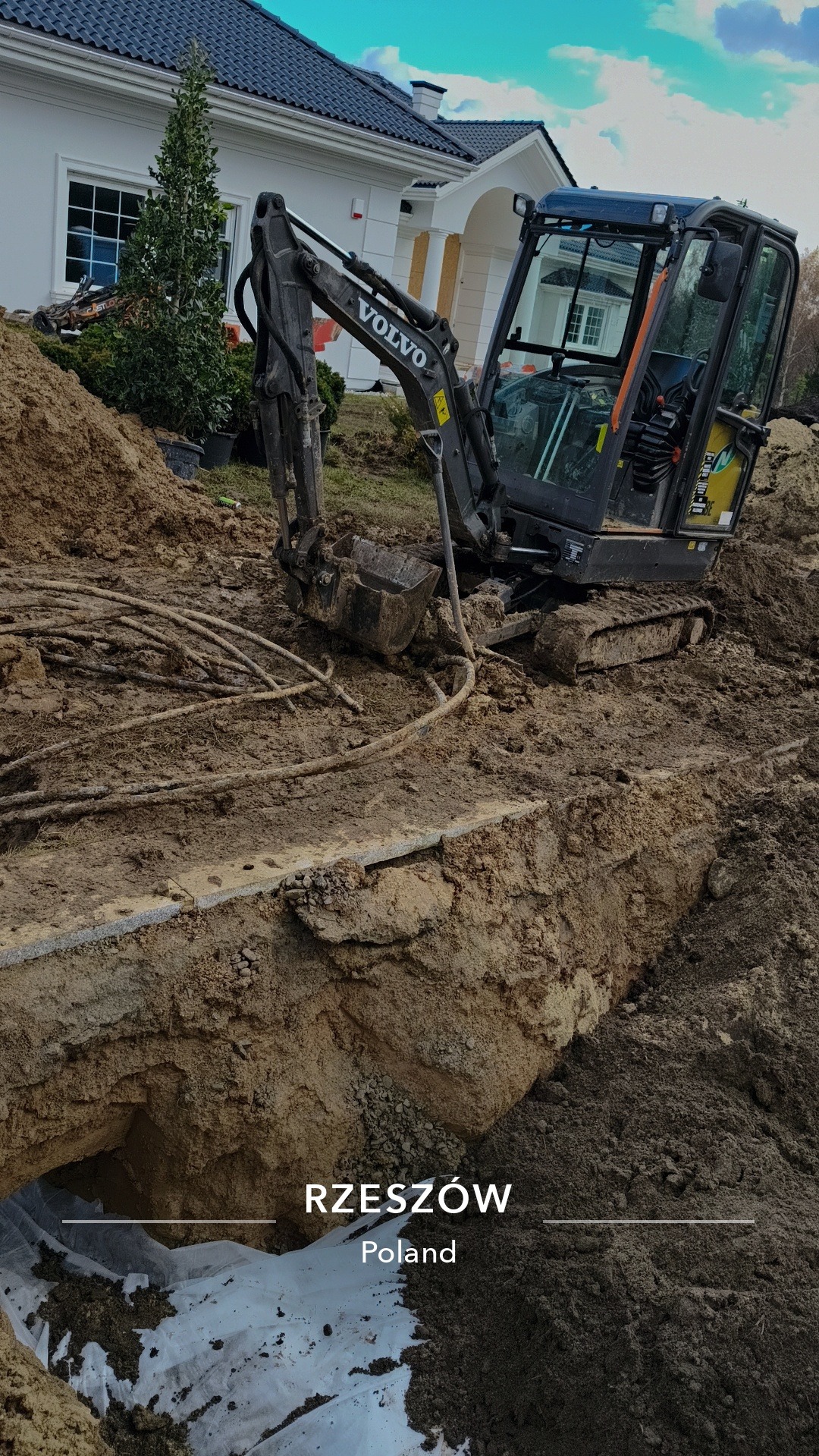 Mini koparka Volvo w wykopie ziemnym na tle domu jednorodzinnego w Rzeszowie. Widoczne kable i folia. Ujęcie z bliska, podkreślające detale wykopu.