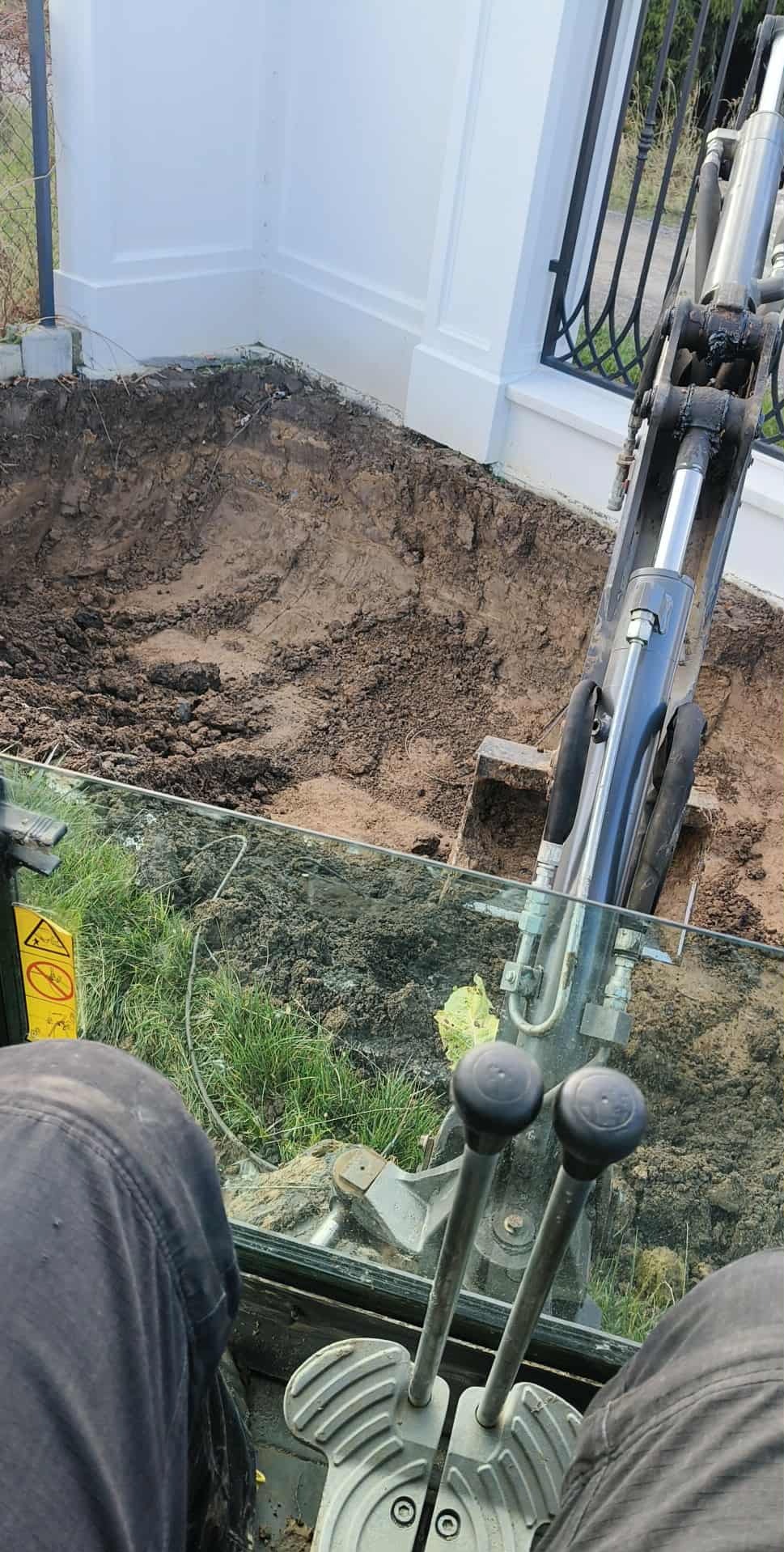 Wykop pod fundament ogrodzenia wykonany minikoparką, widok z kabiny operatora. Świeżo naruszona ziemia przy białym, ozdobnym ogrodzeniu.