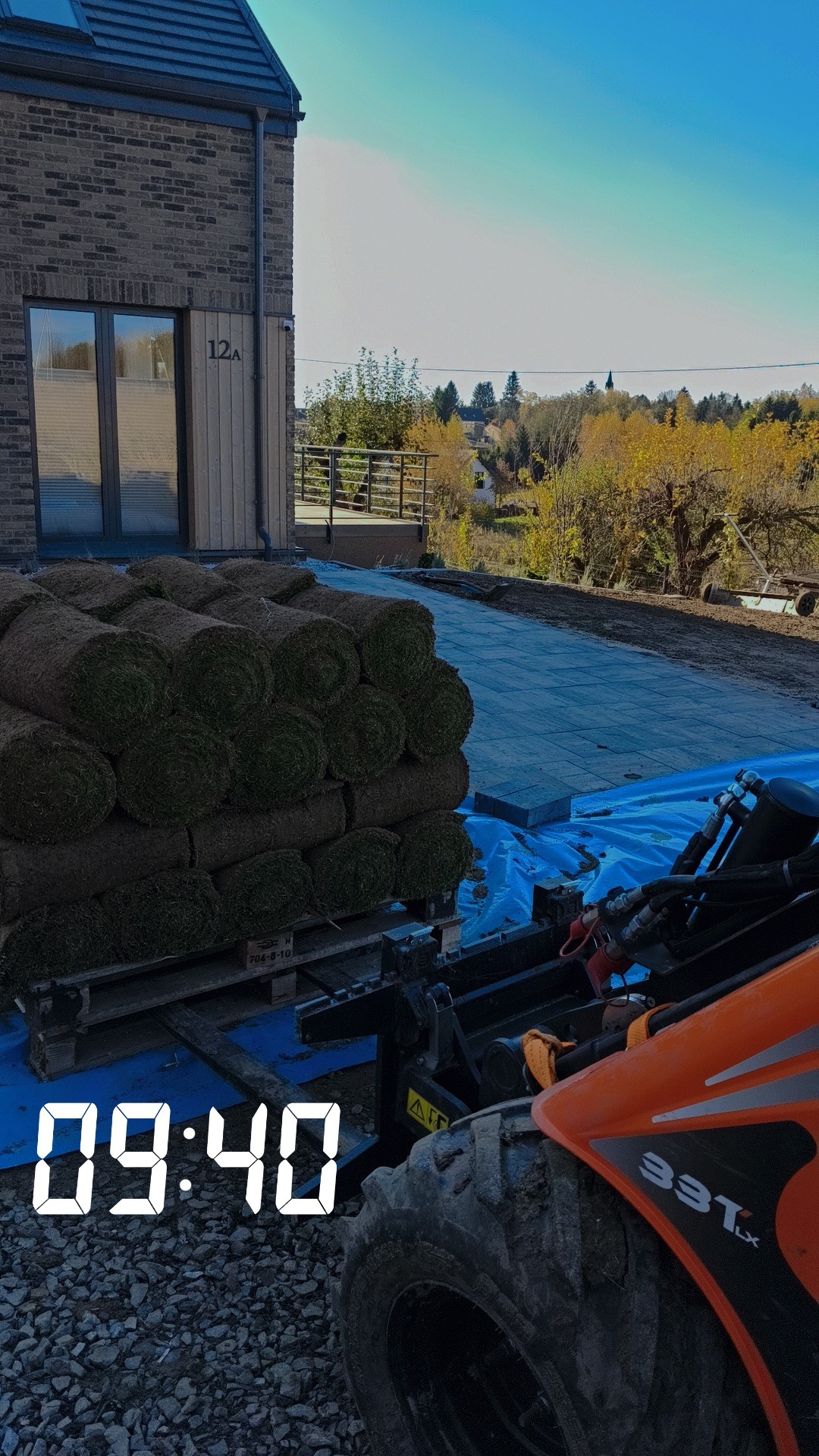 Paleta z rolkami trawy na tle nowo ułożonej kostki brukowej i budynku z numerem 12A, widziana z perspektywy maszyny budowlanej w słoneczny dzień.