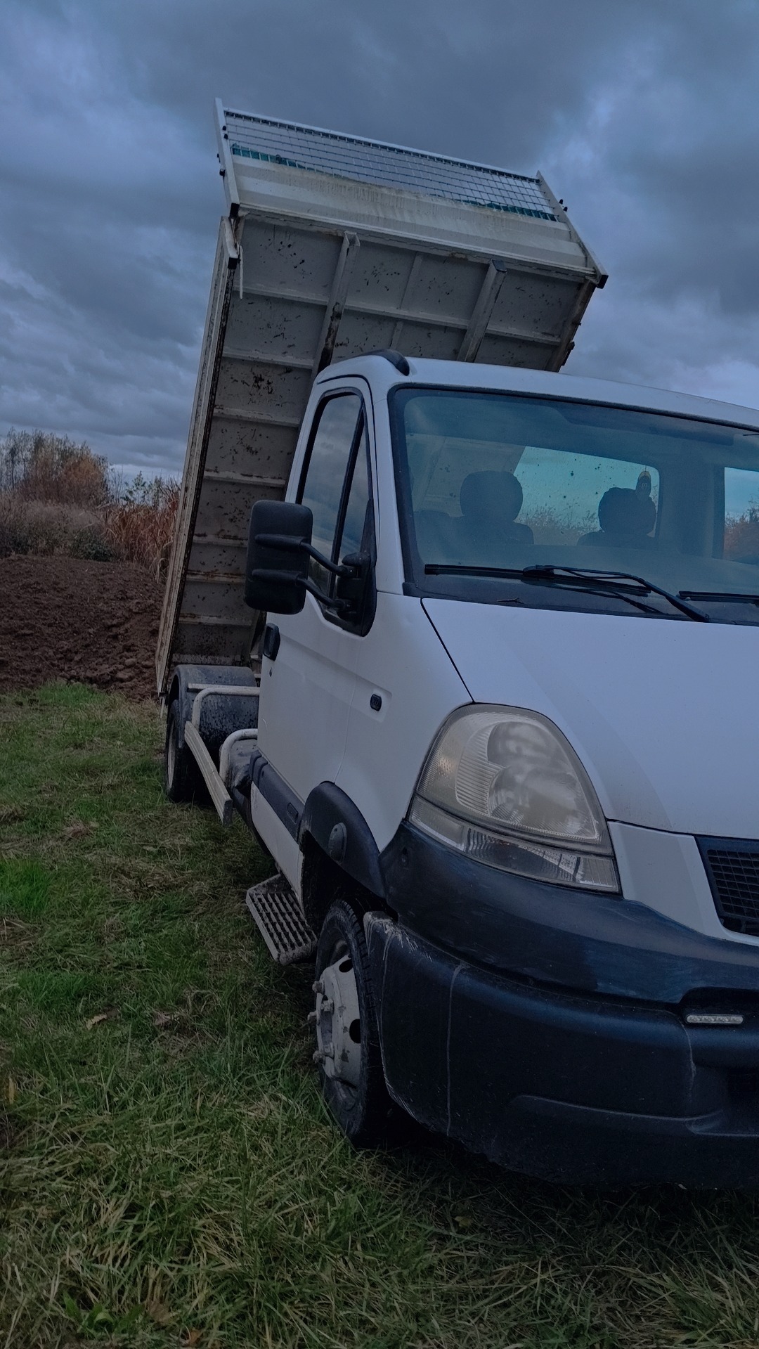 Biały wywrotka z podniesioną skrzynią na tle pochmurnego nieba i zaoranego pola. Widoczne detale: błotniki, koła, reflektory. Ujęcie z dołu, podkreślające wielkość pojazdu.