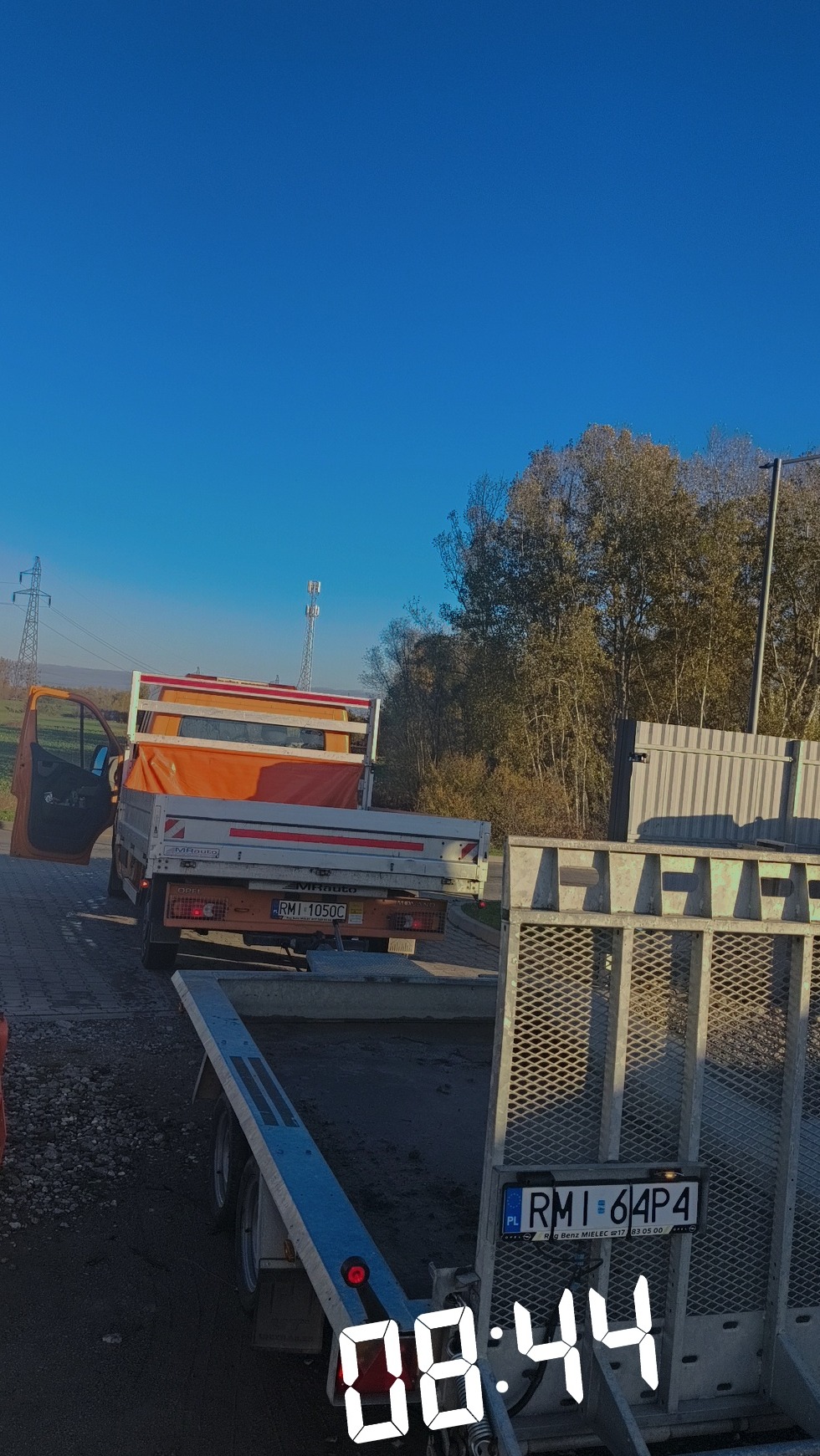 Pomarańczowy samochód dostawczy z otwartymi drzwiami i przyczepą na tle błękitnego nieba i drzew. Widoczny numer rejestracyjny pojazdu i przyczepy.