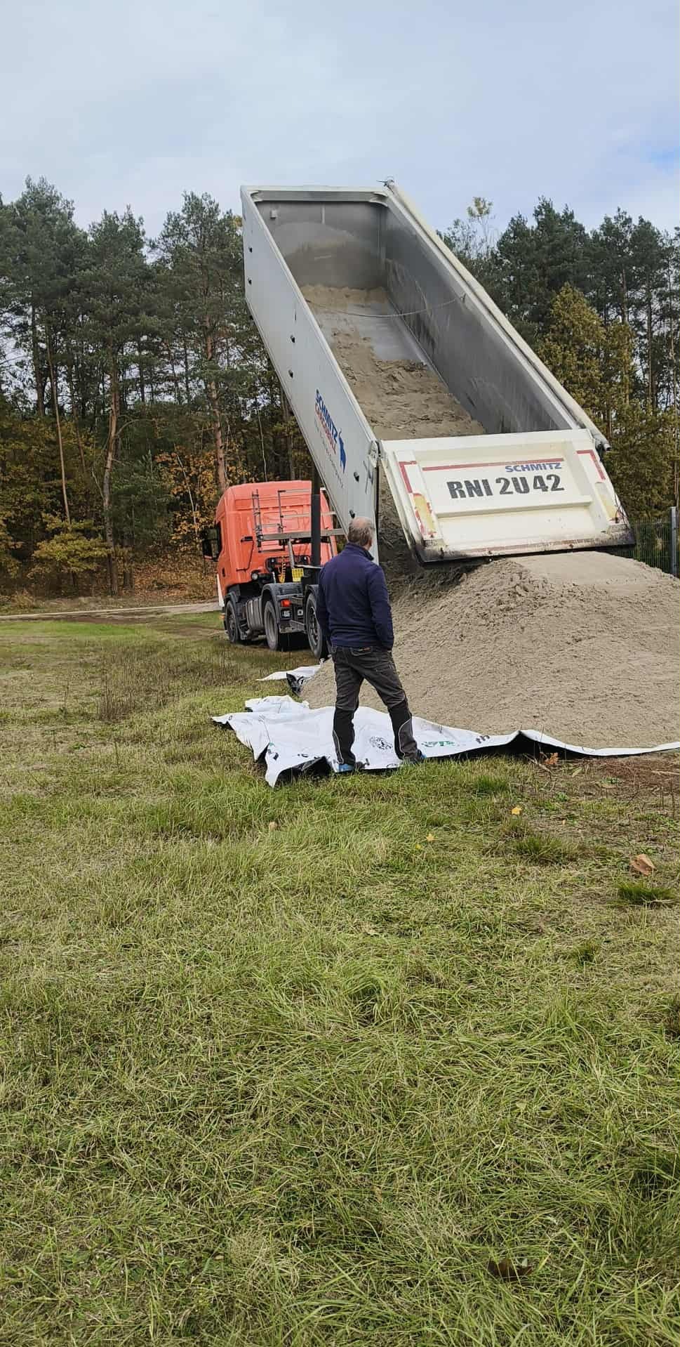 Ciężarówka wyładowuje piasek na trawniku, mężczyzna obserwuje proces. Widoczny las w tle. Wyładunek piasku na przygotowaną plandekę.
