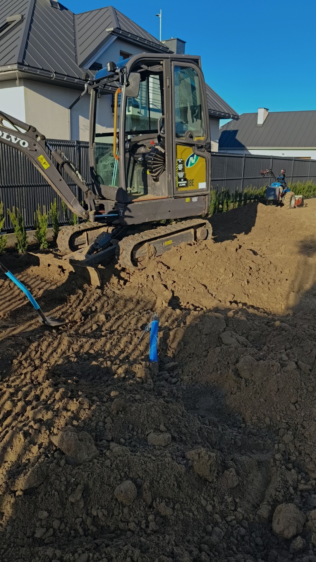 Mini koparka Volvo na placu budowy, obok leży niebieska rura karbowana i łopata. W tle dom jednorodzinny i mały traktorek.