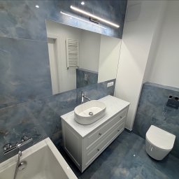 DINO TS Paweł Kwiatkowski - Błękitna łazienka z owalną umywalką nablatową, lustrem z oświetleniem LED i podwieszaną toaletą. Płytki imitujące marmur na ścianach i podłodze.