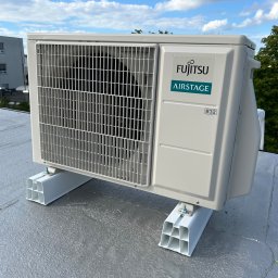 DINO TS Paweł Kwiatkowski - Biała jednostka zewnętrzna klimatyzacji Fujitsu Airstage zamontowana na płaskim dachu, widok z boku, błękitne niebo w tle. Widoczne elementy montażowe.