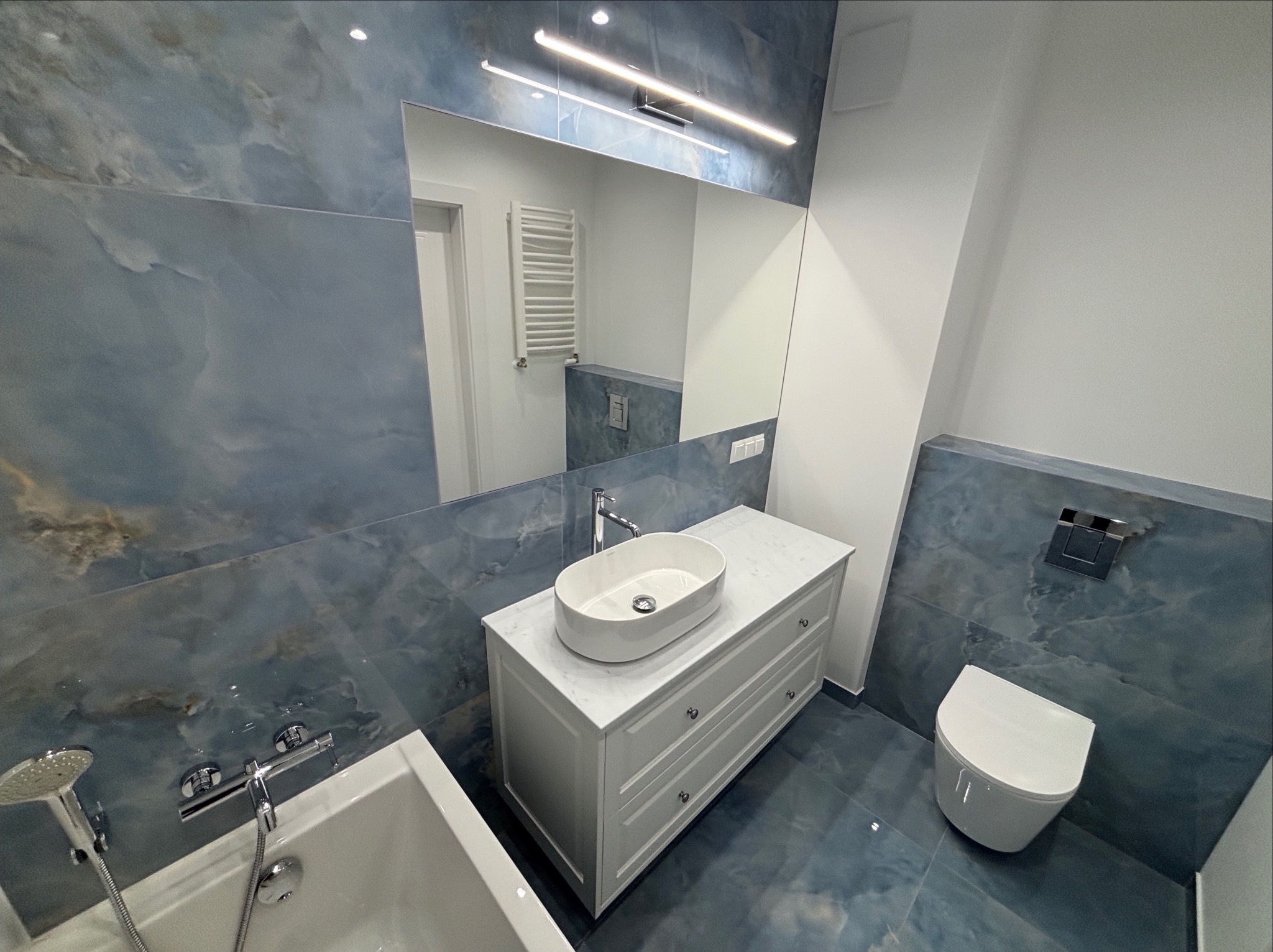 Błękitna łazienka z owalną umywalką nablatową, lustrem z oświetleniem LED i podwieszaną toaletą. Płytki imitujące marmur na ścianach i podłodze.