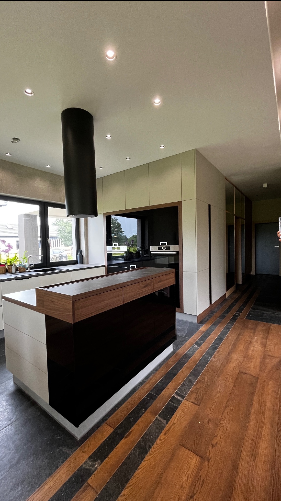 Nowoczesna kuchnia z wyspą, czarnym okapem i drewnianą podłogą z czarnymi wstawkami. Minimalistyczny design, jasne szafki i duże okno.