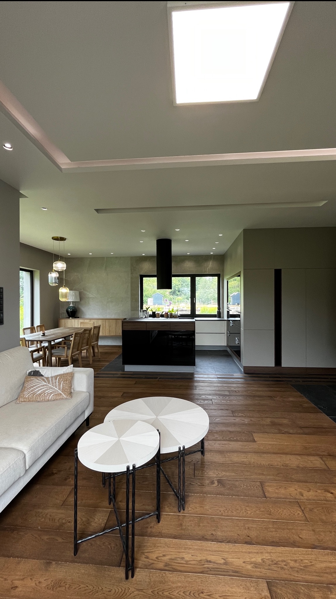 Przestronne wnętrze domu z drewnianą podłogą, białym narożnikiem i okrągłymi stolikami. W tle widoczna kuchnia z czarną wyspą i jadalnia z drewnianym stołem.