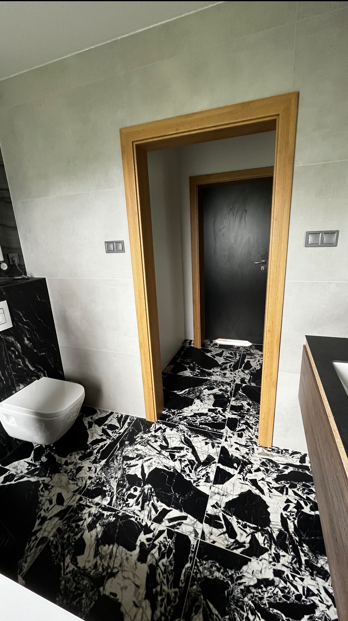 Nowoczesna łazienka z czarno-białą marmurową podłogą i drewnianymi futrynami drzwi, widok na kolejne pomieszczenie. Minimalistyczny design.