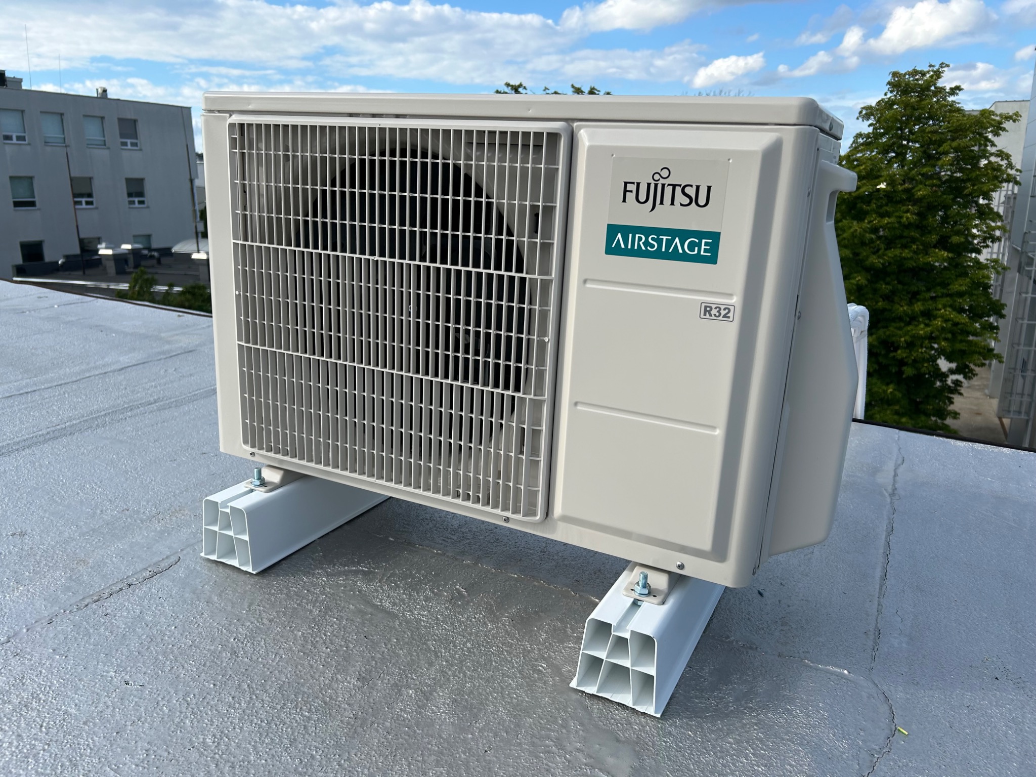 Biała jednostka zewnętrzna klimatyzacji Fujitsu Airstage zamontowana na płaskim dachu, widok z boku, błękitne niebo w tle. Widoczne elementy montażowe.