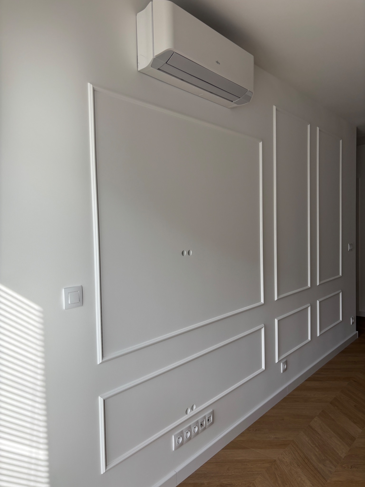 Biała ściana z dekoracyjnymi listwami, gniazdkami i klimatyzatorem Fujitsu. Parkiet w jodełkę na podłodze. Promienie słońca na ścianie.