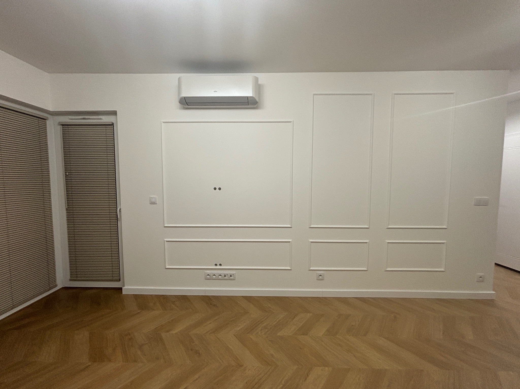 Eleganckie wykończenie wnętrza: białe ściany z dekoracyjnymi panelami, parkiet w jodełkę i klimatyzator. Widoczne gniazdka elektryczne i rolety w oknie.