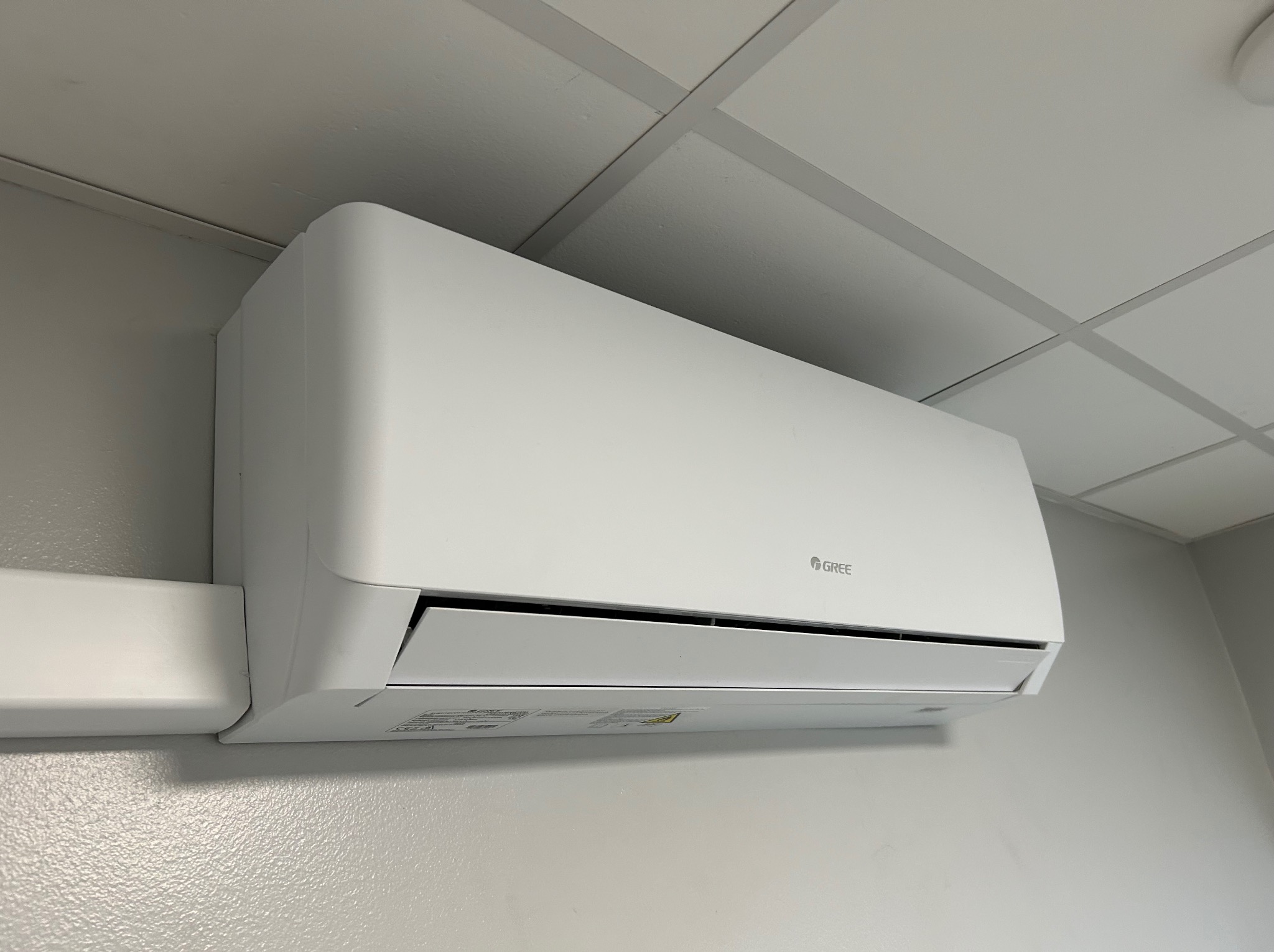 Biała klimatyzacja Gree zamontowana na jasnej ścianie pod sufitem kasetonowym. Widoczny fragment listwy przypodłogowej. Minimalistyczny design.