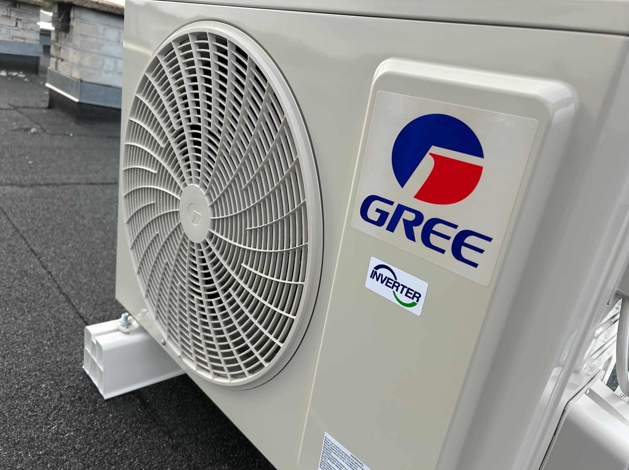 Zewnętrzna jednostka klimatyzacji Gree Inverter na dachu, widoczny wentylator i logo marki. Szara, gumowa powierzchnia dachu w tle.