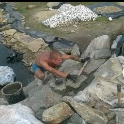 Visual Garden 5.3.0.9.0.8.7.2.8 - Budowa oczka wodnego: mężczyzna w rękawicach układa kamienie brzegowe, w tle woda, kamień ozdobny i folia. Widoczne narzędzia: poziomica i młotek.