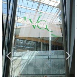 Visual Garden 5.3.0.9.0.8.7.2.8 - Montaż kamiennej ściany wewnątrz budynku z przeszklonym dachem. Dwóch pracowników na rusztowaniach, w pomarańczowych kamizelkach, układa kamień. Widoczne narzędzia i materiały.