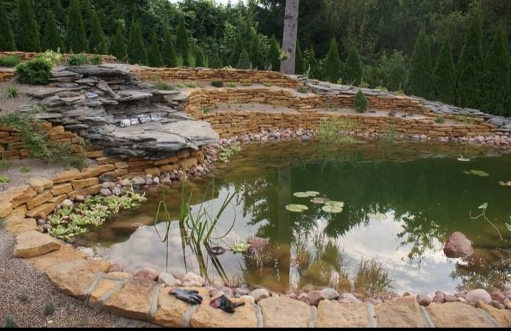 Oczko wodne z kamienną kaskadą i obrzeżem z piaskowca, otoczone zielenią. Na pierwszym planie leżą robocze rękawice. Refleksy drzew w tafli wody.