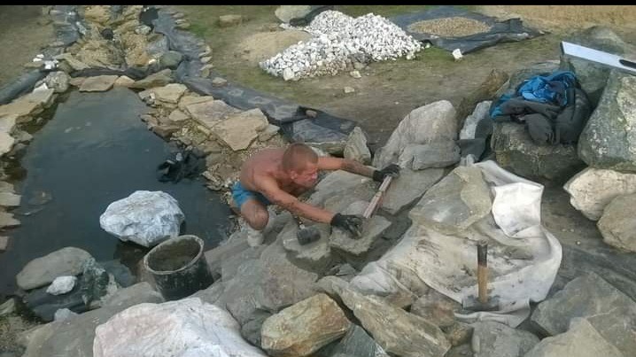 Budowa oczka wodnego: mężczyzna w rękawicach układa kamienie brzegowe, w tle woda, kamień ozdobny i folia. Widoczne narzędzia: poziomica i młotek.