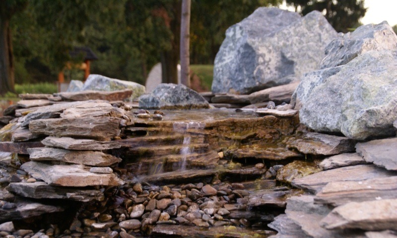 Ogród skalny z kaskadą wodną, zbudowany z płaskich kamieni i otoczaków, z zielenią w tle. Element małej architektury ogrodowej.