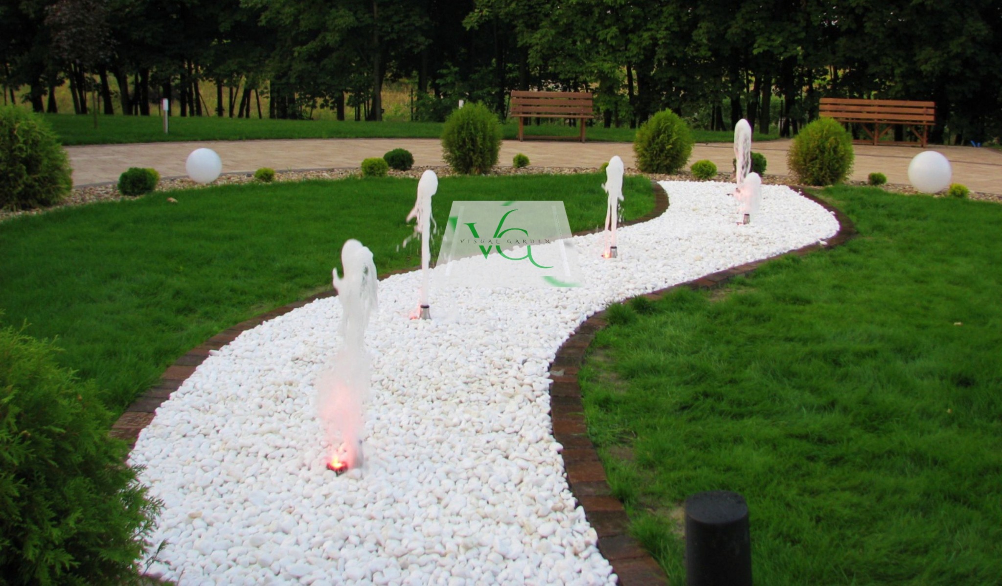 Ogród z ścieżką z białych kamieni, obrzeżem z cegły i fontannami. W tle trawnik, krzewy i ławki. Na ścieżce logo firmy Visual Garden.
