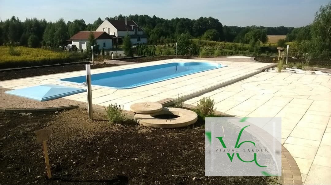 Kąpielowy basen z jasną kostką brukową na tarasie, otoczony zielenią i domem w tle. Widoczne studzienki kanalizacyjne i oświetlenie.