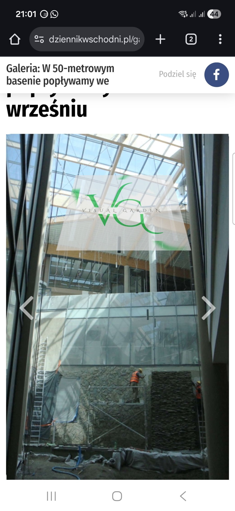 Montaż kamiennej ściany wewnątrz budynku z przeszklonym dachem. Dwóch pracowników na rusztowaniach, w pomarańczowych kamizelkach, układa kamień. Widoczne narzędzia i materiały.