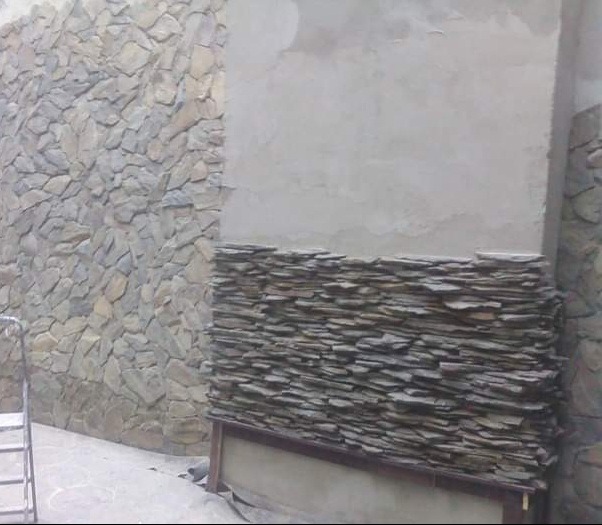 Fragment ściany z częściowo ułożonym kamieniem dekoracyjnym, widoczna drabina i surowy tynk. Prace wykończeniowe na zewnątrz budynku.