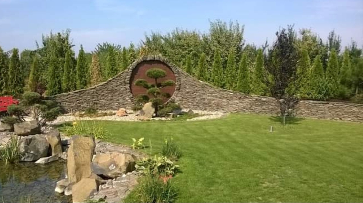 Ogród z kamienną ścianą i okrągłym oknem, w którym rośnie bonsai. W tle wysokie drzewa iglaste, z przodu trawnik i oczko wodne z kamieniami.