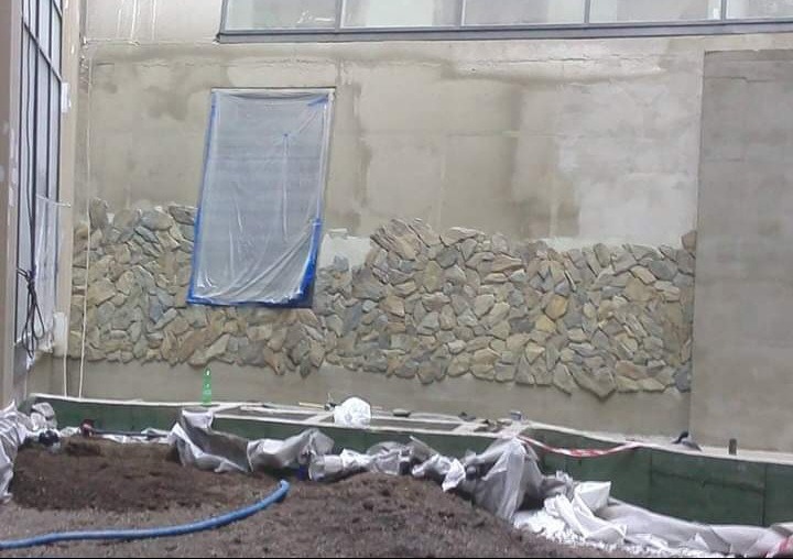 Wnętrze w trakcie prac: częściowo wykończona ściana z kamienia, zabezpieczone okno folią, ziemia i narzędzia widoczne na pierwszym planie.