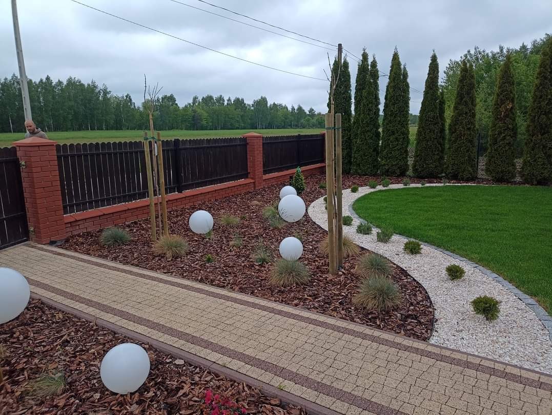 Ogród po realizacji: ścieżka z kostki, trawnik z rolki, ozdobne krzewy i drzewa iglaste, kora, białe kule oświetleniowe, ogrodzenie z ceglanymi słupkami.