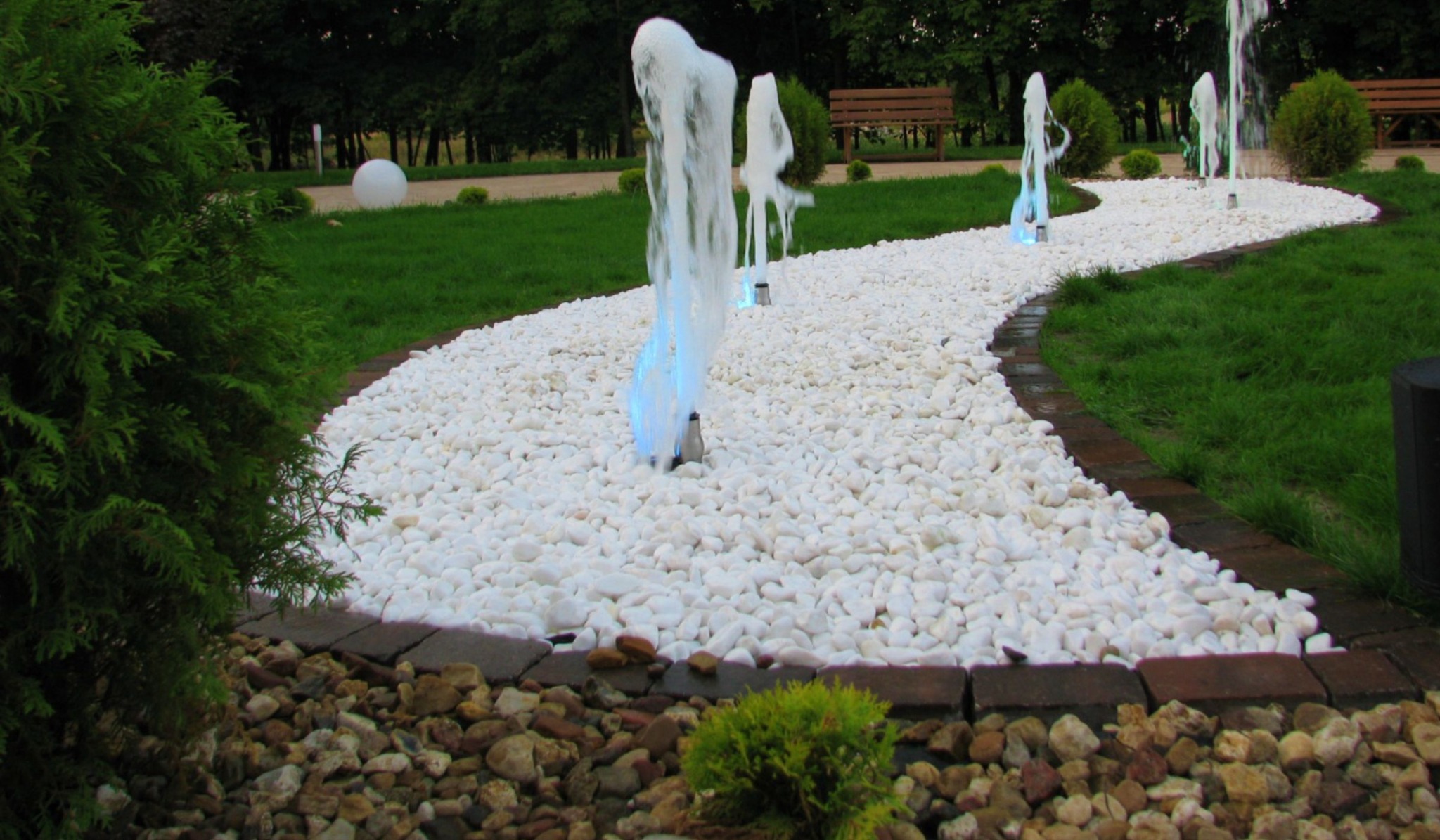 Ogród z białym żwirem i fontannami, obrzeża z kostki brukowej, zieleń trawników i krzewów. Elementy małej architektury parkowej w tle.