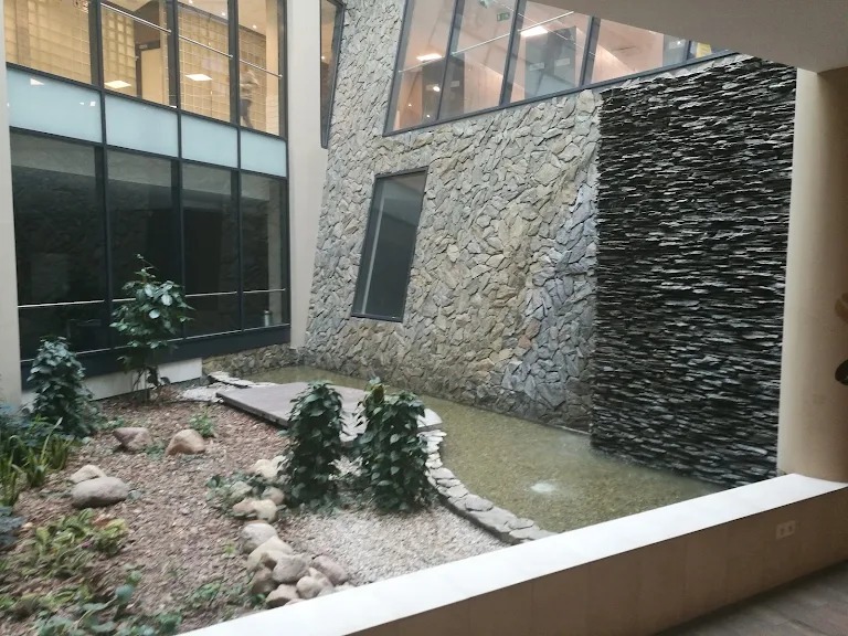 Wewnętrzny dziedziniec z kamienną ścianą, wodą i roślinnością. Widoczne okna biurowe i elementy architektoniczne. Dekoracyjny ogród wewnątrz budynku.
