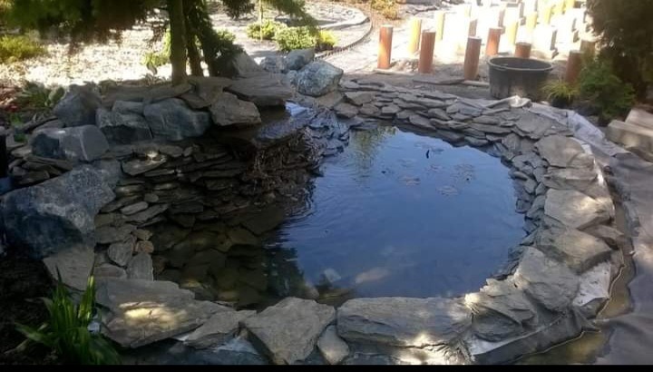 Oczko wodne z kamienną obudową, otoczone zielenią i elementami dekoracyjnymi. Widoczne kamienie różnej wielkości tworzące kaskadę i obrzeże zbiornika.